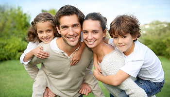 A smiling family of four with two children outdoors on a grassy lawn